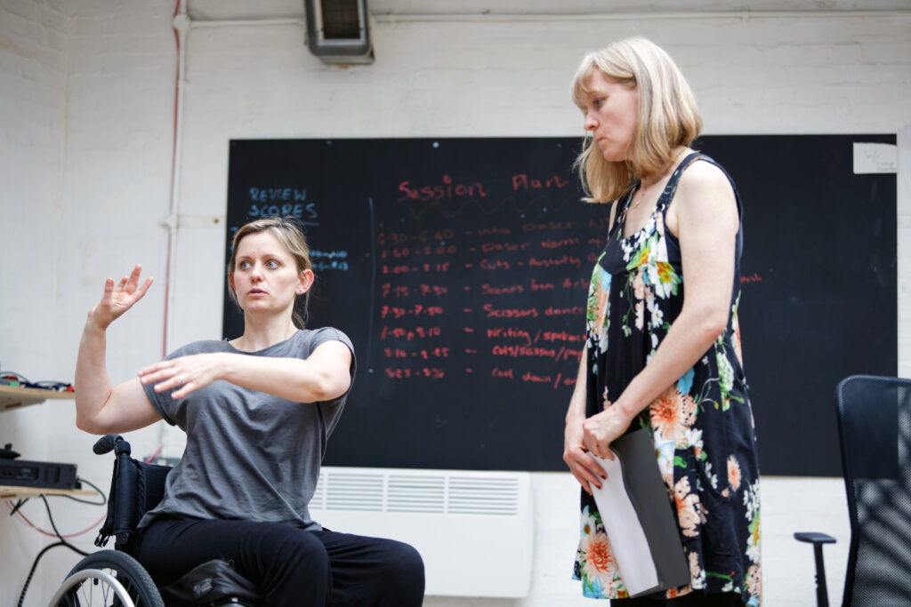 Two blonde women in conversation, one standing, one seated in a wheelchair.