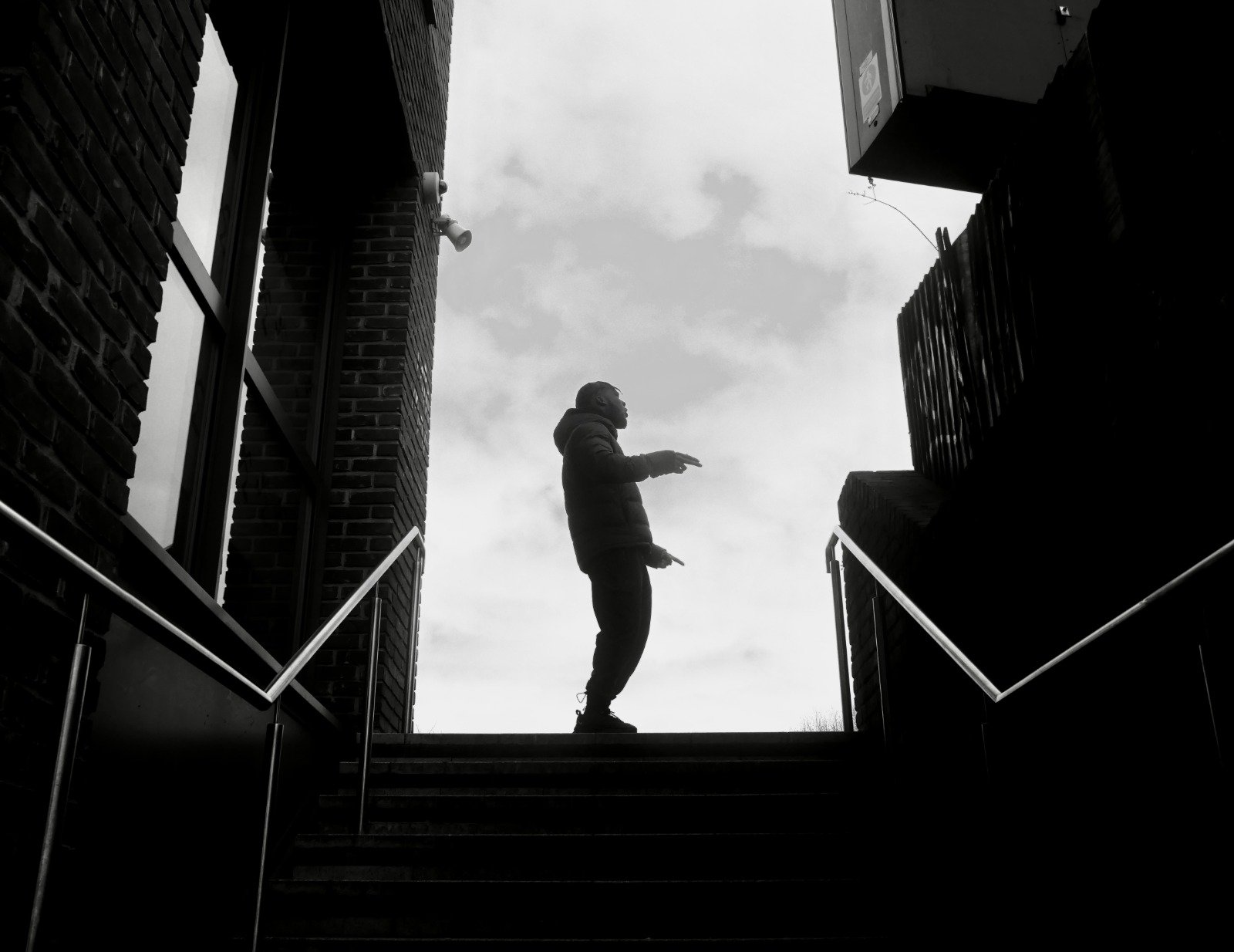 A Black man talking expressively at the top of the stairs in a new estate, black and white filter