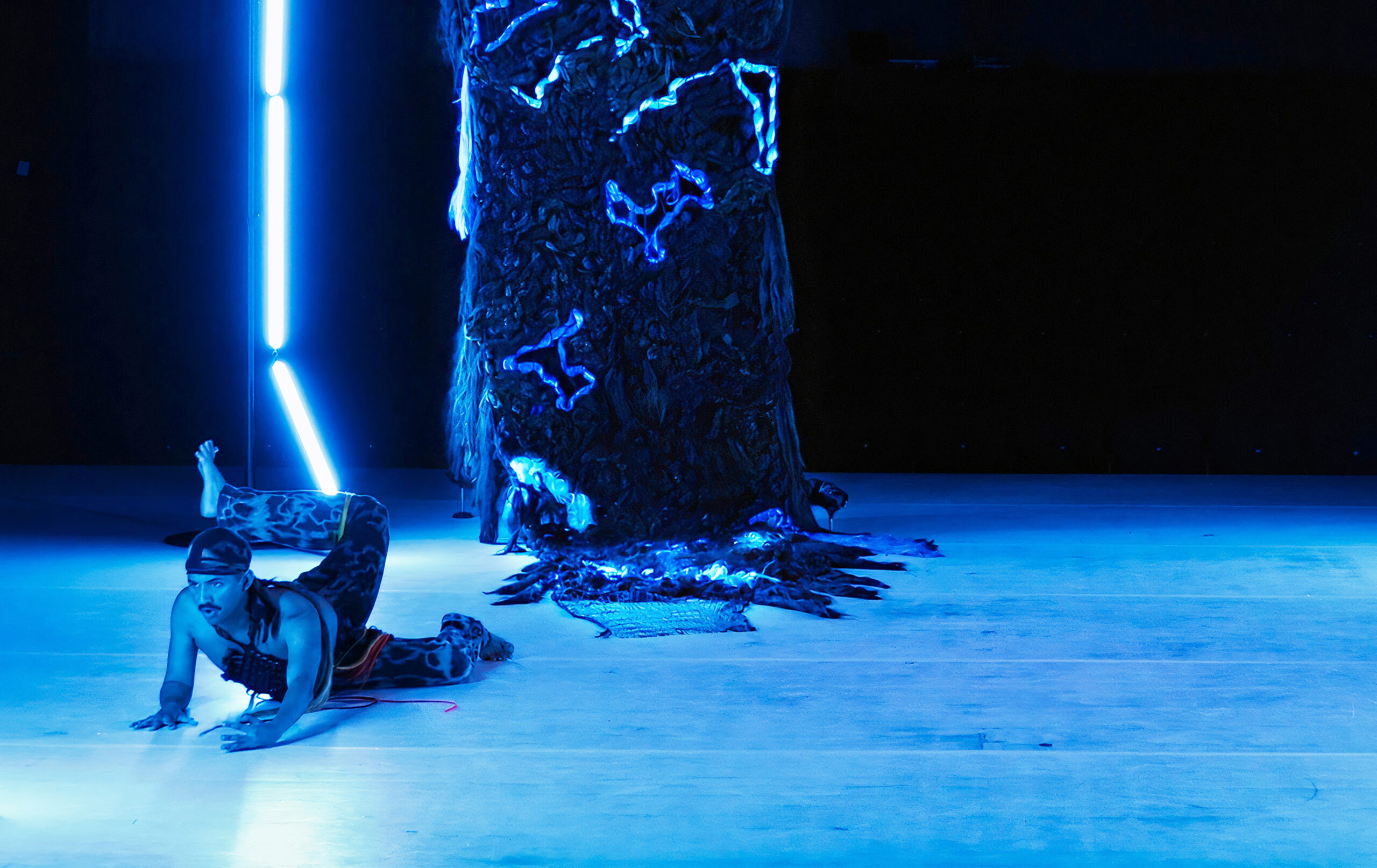 Performer Adam is lying with their belly to the floor. Their head is raised and one leg in patterned trousers is raised behind them. Their foot is visible above their head and their toes are splayed. The lighting is casting a blue hue onto Adam and the giant sculpture of a weave behind them.