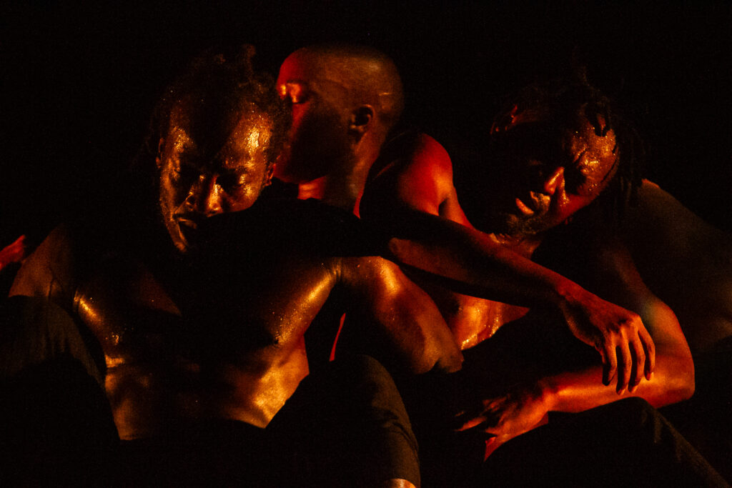 Theophilus Alade, Nosiphiwo Samente and Bafana Matea dancing aginst each other in a dark setting with red lighting on them. Cane Warriors cast and performance images ©2023 Richard Budd