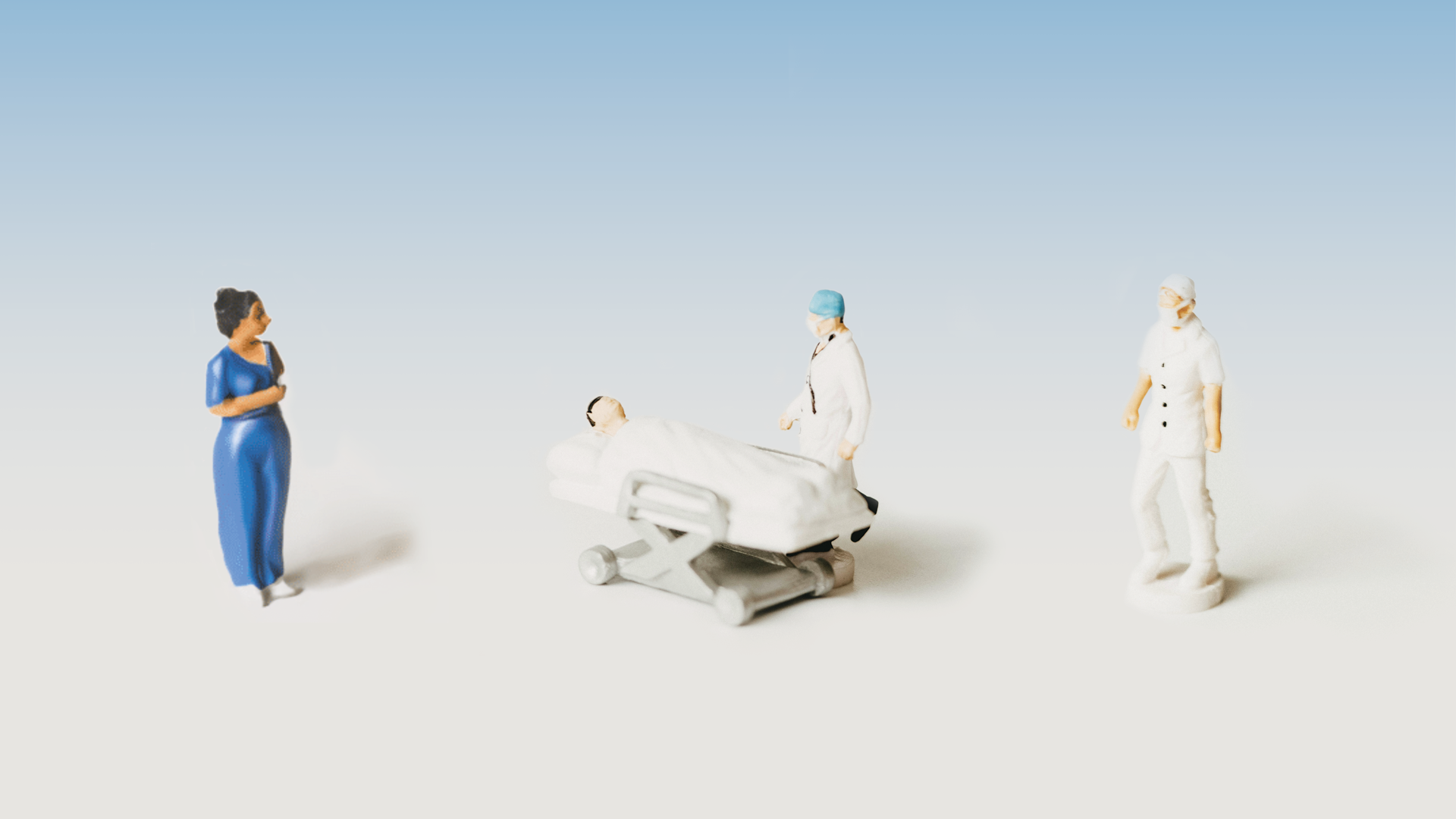 Four Minifigures are positioned in front of a blue-gradient background. They are nurses in scrubs. The figures on the far left and right are looking caringly over patients in their beds. The two central figures stand alone.