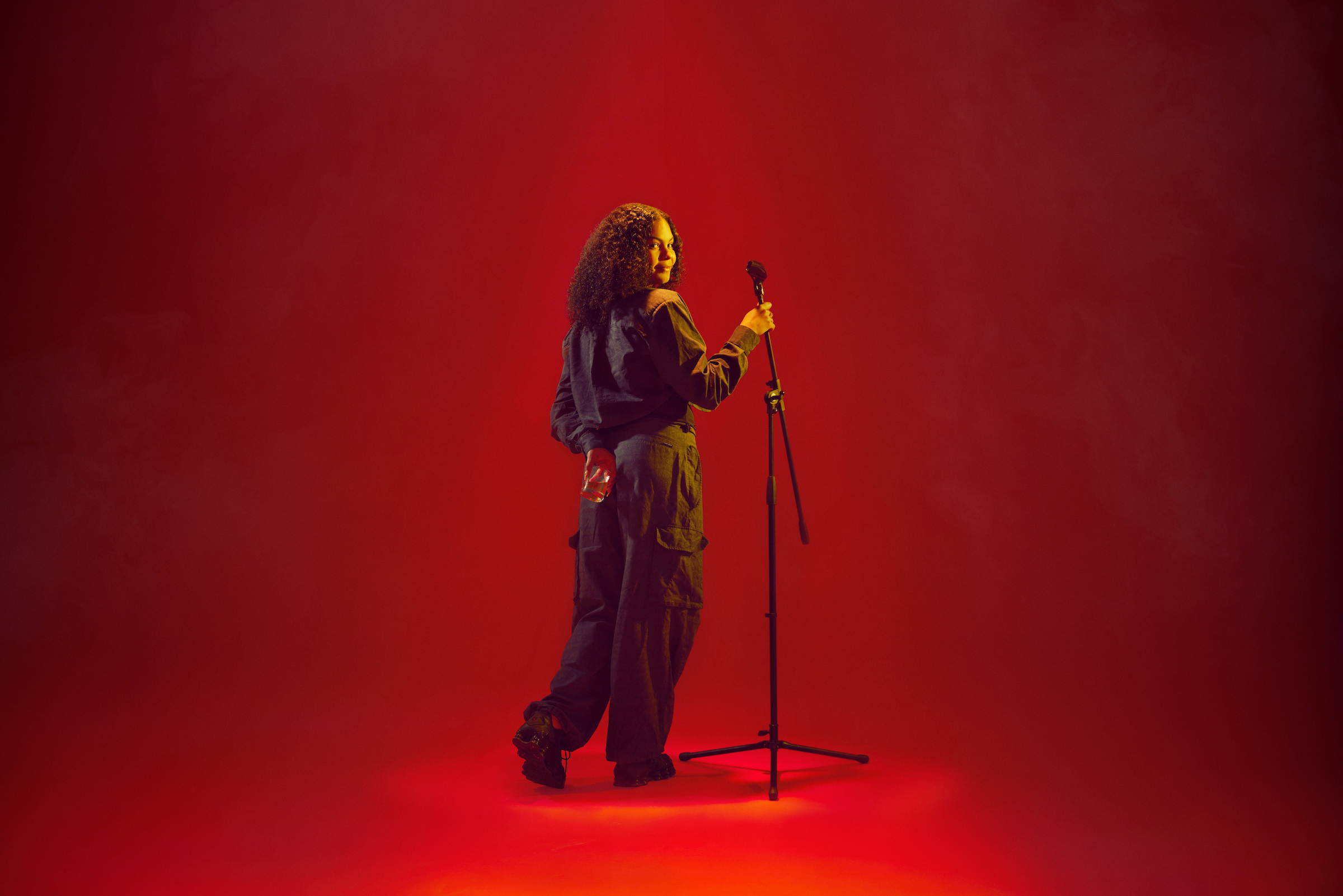 India Wilson as Hennessy Jade, turning back to the camera with a mic stand and a glass of alcohol. Photo by Rich Lakos