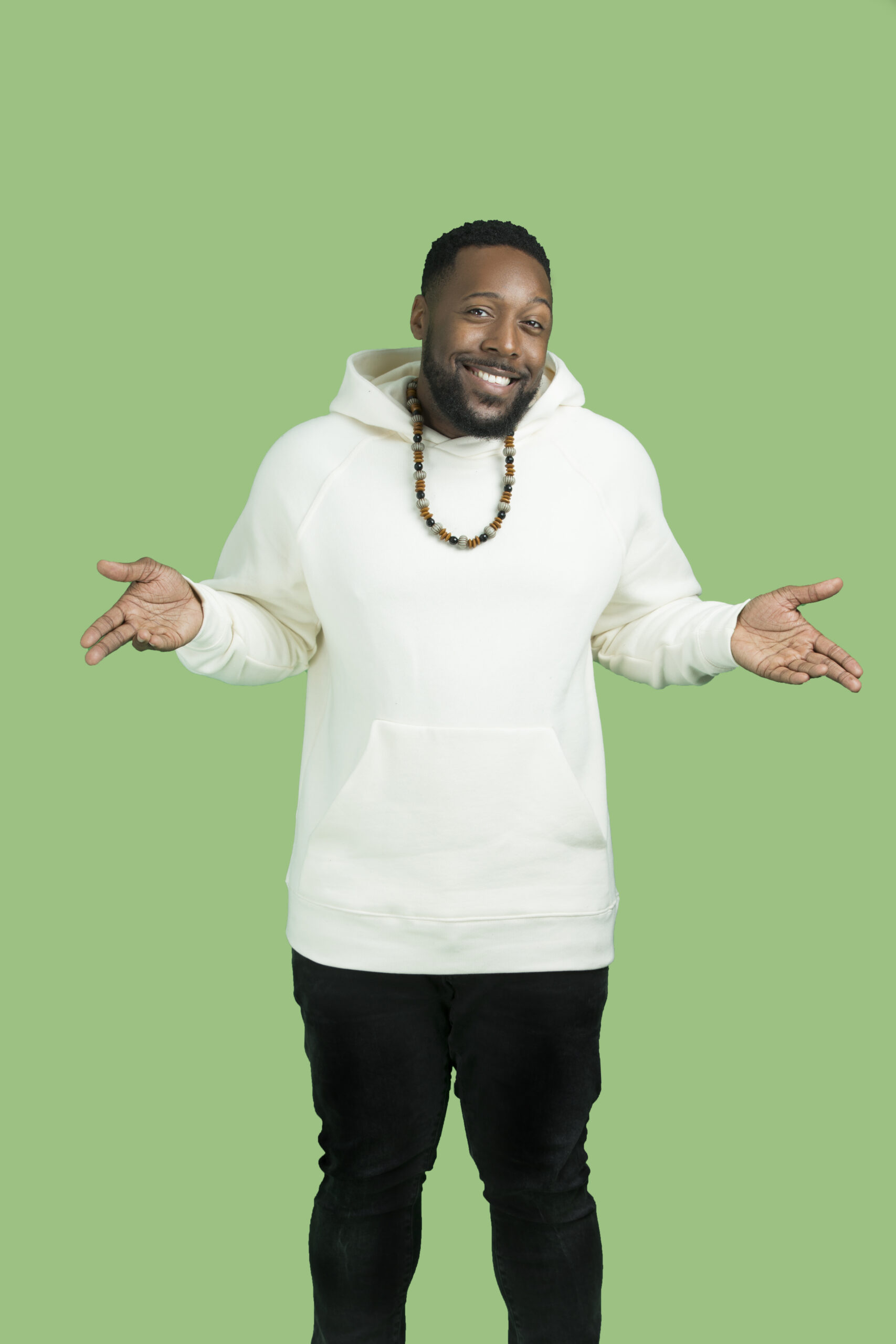 A man smiling and shrugging with his hands out to the sides. He is wearing a white hoodie, a beaded necklace, and black pants, standing against a plain green background