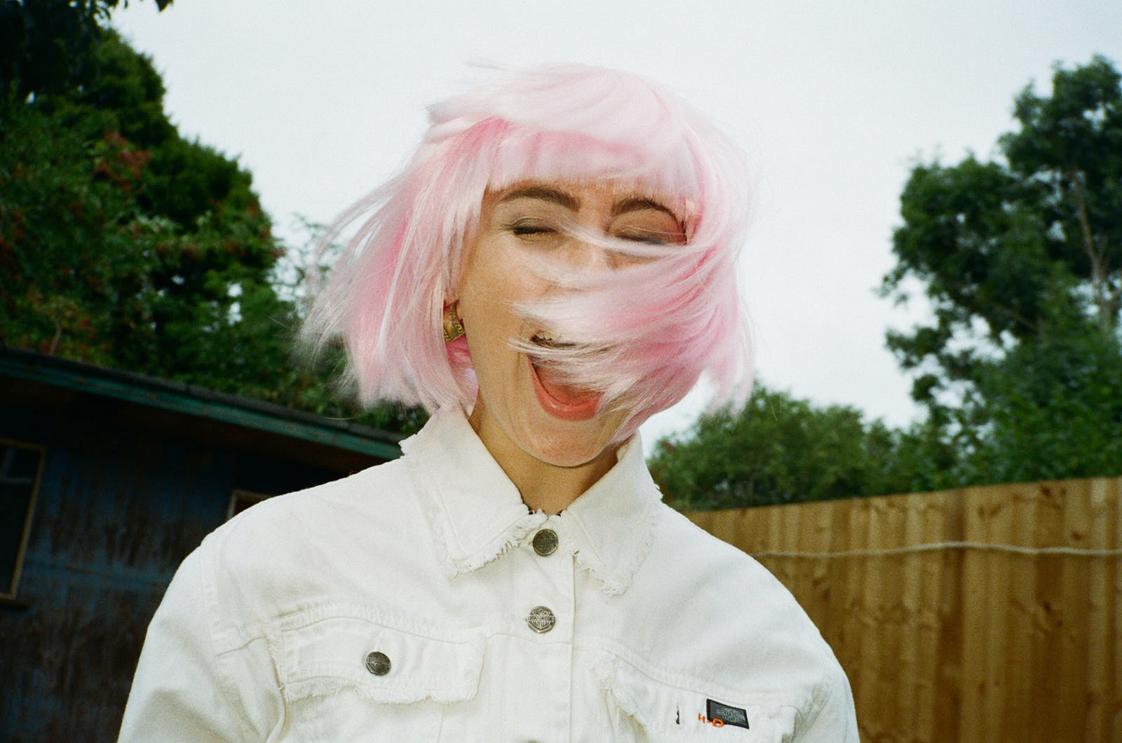 A young white woman in a pink wig and white jacket. Her mouth is open as if screaming with joy. As she shakes her head the pink hair covers half of her face.