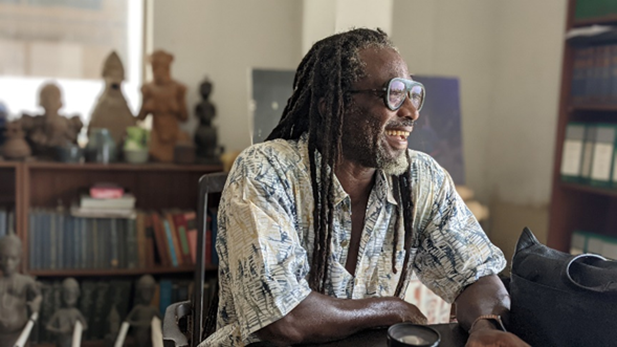 A photograph of the writer Biyi Bándélé sitting at a table, relaxed and laughing.