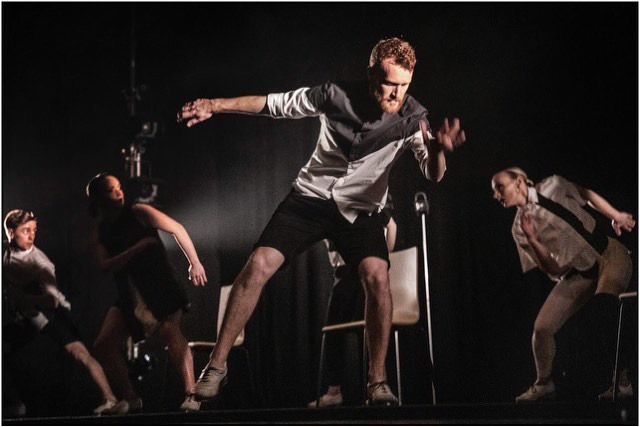 A White man tap dancing wuth his arms in movement, behind him is an ensemble of other dancers