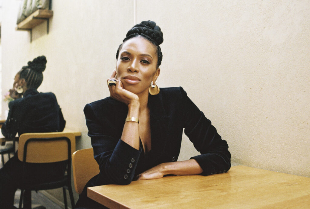 A woman seated at a wooden table, resting her chin on her hand with a confident expression. Her hair is styled in a braided updo, and she wears gold hoop earrings, a black blazer, and subtle makeup. A mirror behind her reflects part of her profile and the back of a chair.