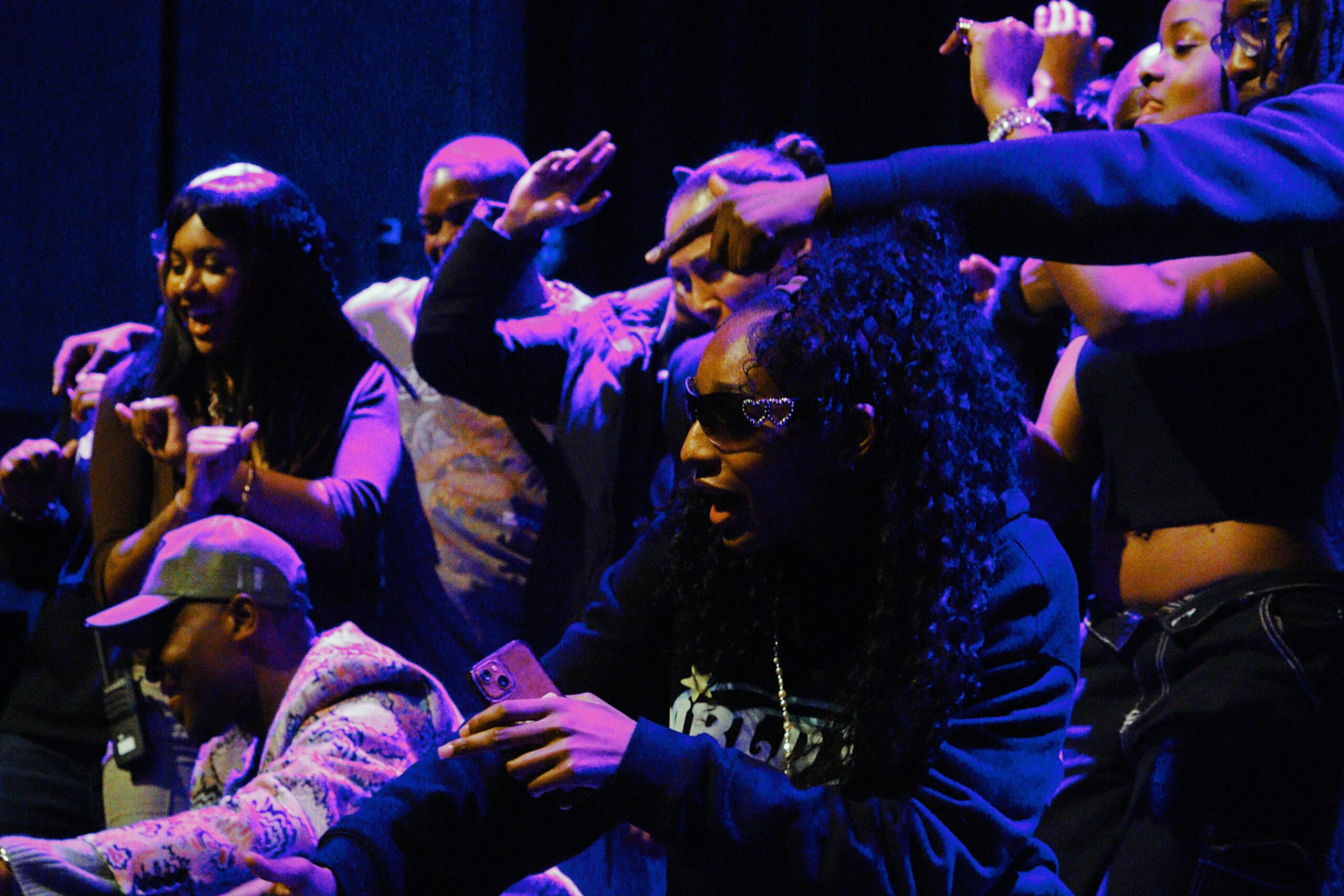 A lively group of young people dancing and celebrating together on stage under colourful lighting. One woman in the foreground wears heart-shaped sunglasses and holds a phone, while others around her raise their hands and smile, capturing the energy and joy of the moment.