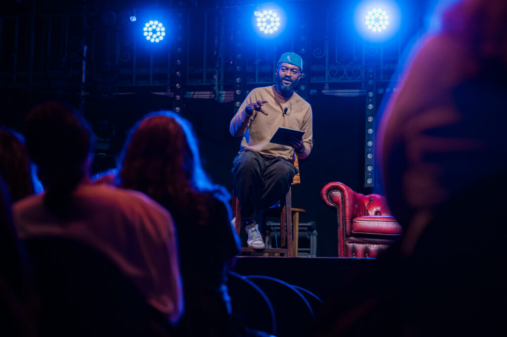 A bearded man of African ethnicity sits on a barstool facing the audience. He wears a brimless green hat and holds a tablet. There is a red leather sofa behind him.