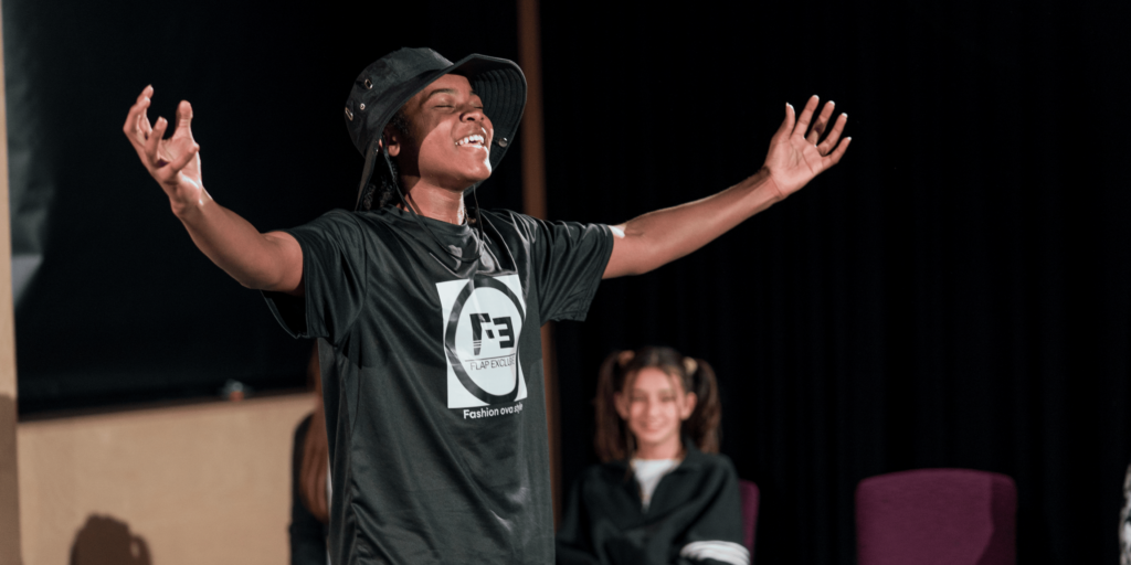 A young Black boy performing on stage. His eyes are closed and both arms outstretched. 